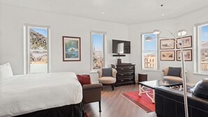 A modern hotel room with a large bed, a sofa, a coffee table, and a TV. There are multiple windows providing views of the outdoors. - Independence Square Unit 303 | Corner Studio with Premier Finishes, Downtown Location (Aspen)