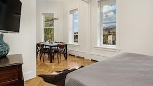Ein Schlafzimmer mit Bett, Fernseher, Essecke mit Stühlen und Tisch sowie großen Fenstern mit Blick ins Freie.