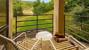 A balcony with a view of a forested area and a dirt path. There are two chairs and a small table.