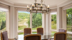 A dining area with a glass table surrounded by striped chairs, a chandelier, and large windows offering a view of a forested landscape.