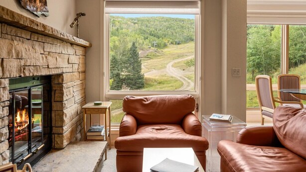 A cozy living room with a stone fireplace, leather armchair, and a view of a forested area through a large window.