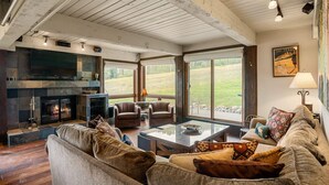 Un salon chaleureux avec une cheminée, des sièges confortables et de grandes fenêtres donnant sur l’extérieur.