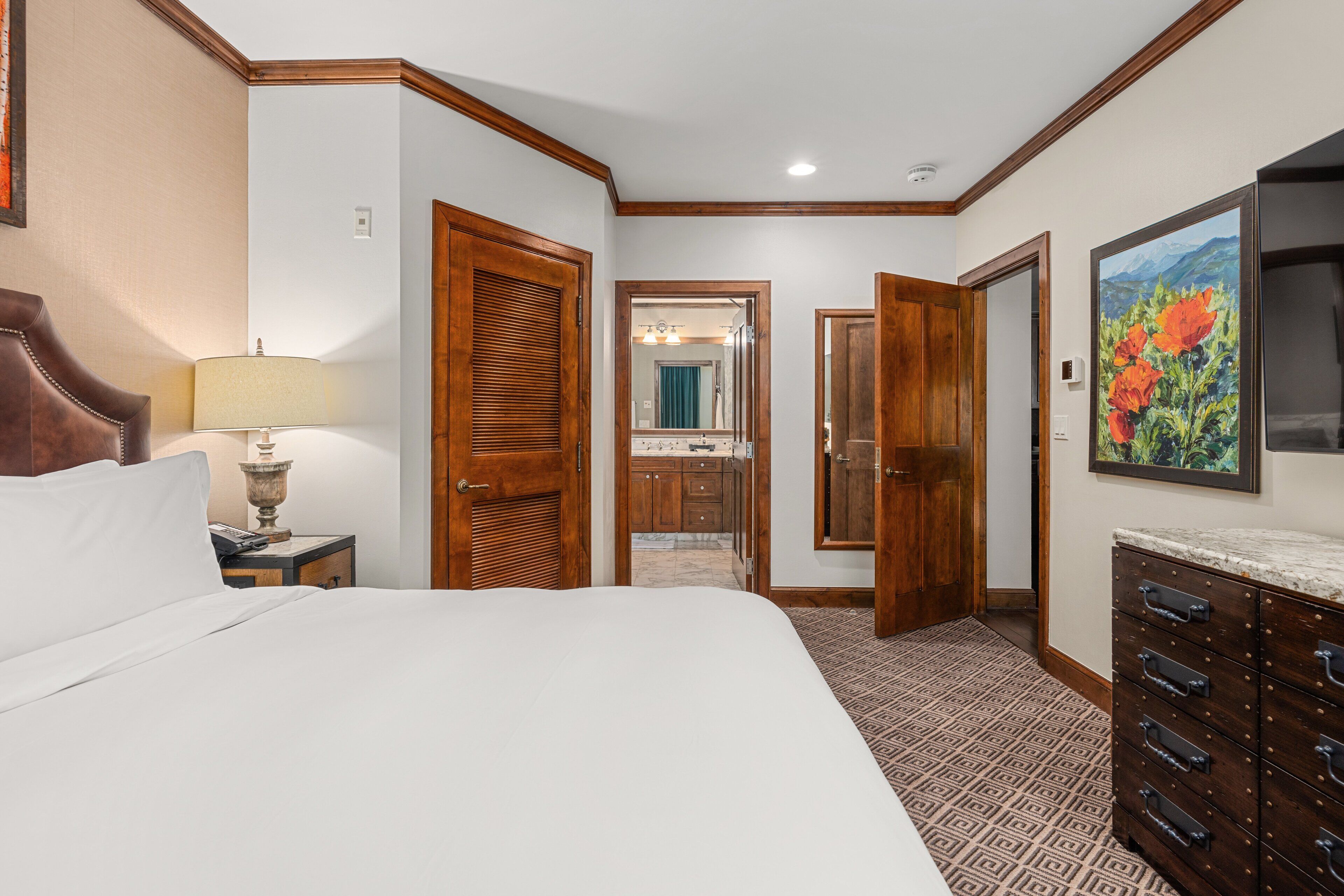 Une chambre d’hôtel avec un grand lit, des portes en bois, une salle de bain visible à travers une porte ouverte et une commode avec des tiroirs.