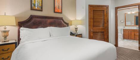 A hotel room with a large bed, two bedside lamps, a wooden headboard, a nightstand, a dresser, and a bathroom visible through an open door.