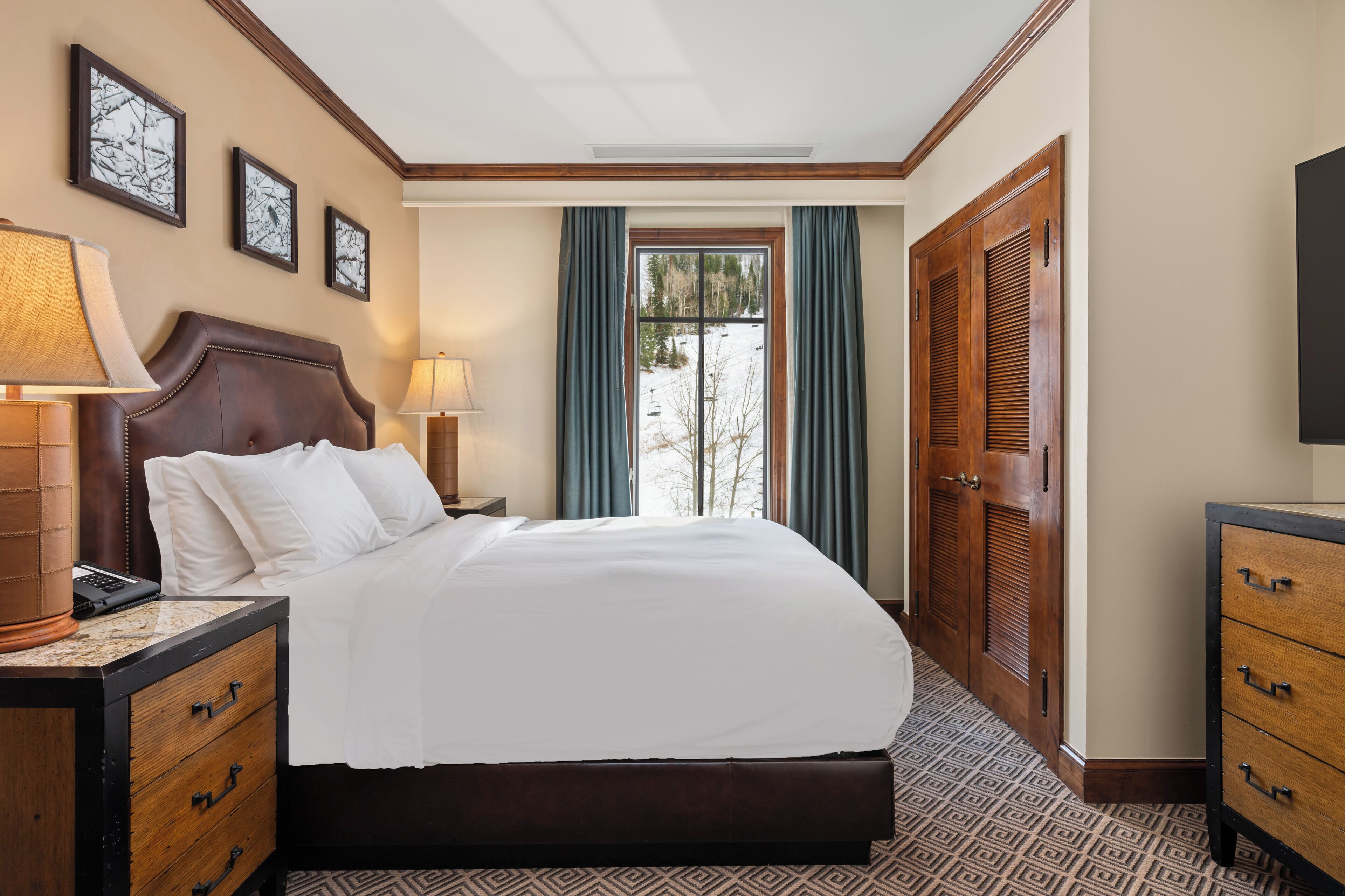 A hotel room with a large bed, wooden headboard, two bedside tables with lamps, a dresser, and a window with curtains.