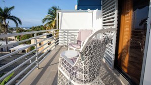 A balcony with a white wicker chair, a small table, and a view of the ocean and palm trees. - MB Apartment A (Puerto Plata)