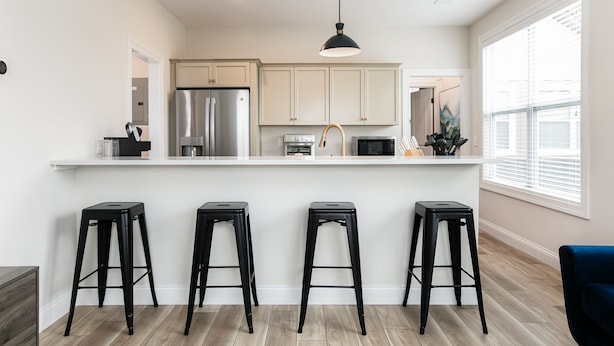 A modern kitchen with white cabinets, a stainless steel refrigerator, a microwave, and a black pendant light. There are four black stools at the counter. - Victory Views Estate: Luxury Guest Loft (South Bend)