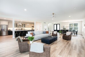A spacious living room with a sofa, ottoman, and chairs, a kitchen in the background, and a dining area with a table and chairs.
