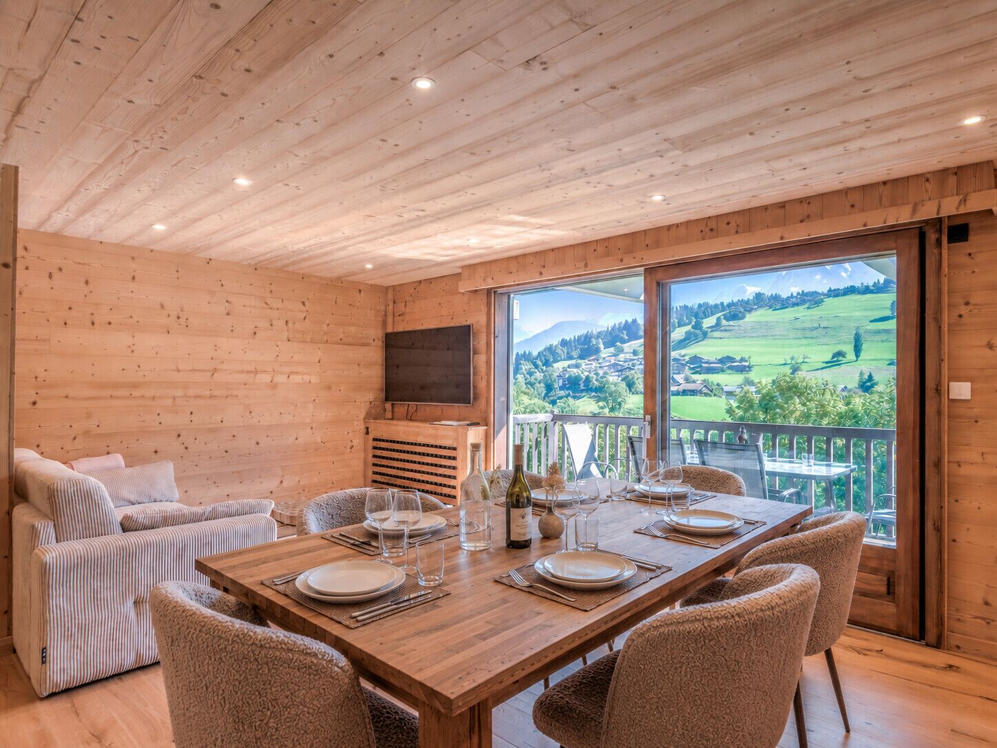 Un interior de madera con una mesa de comedor preparada para seis personas, un sofá y un televisor montado en la pared.