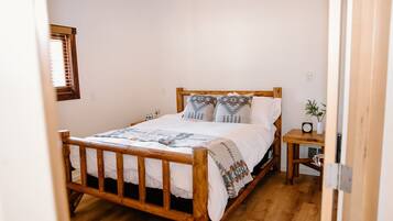 A wooden bed with a headboard, a bedside table with a clock, and a window with blinds.