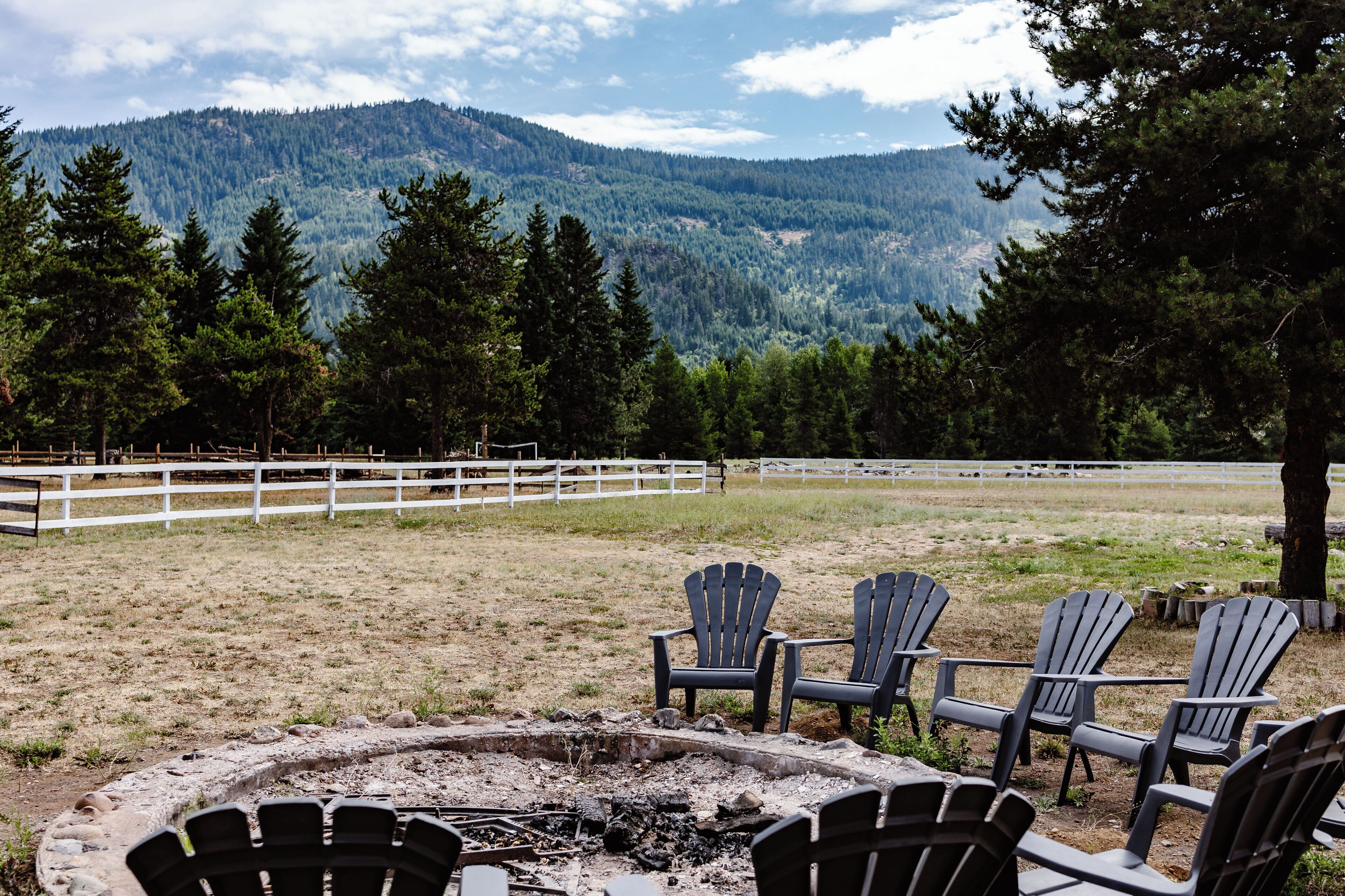 A scenic outdoor area with a fire pit, Adirondack chairs, and a white fence.