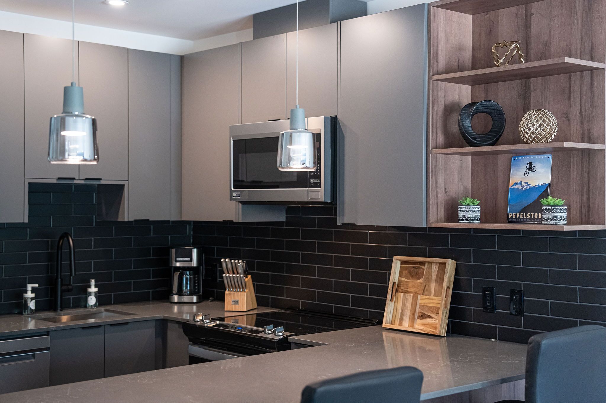 A modern kitchen with dark tiled backsplash, stainless steel appliances, and wooden shelves with decorative items.