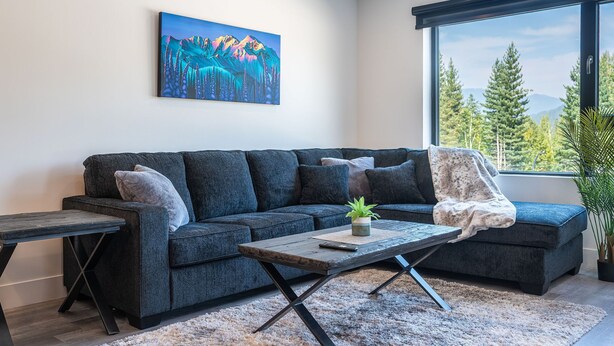 A modern living room with a dark grey sectional sofa, a wooden coffee table, and a large window offering a view of trees and mountains. - Alpine Haven by Revelstoke Vacations (Revelstoke)