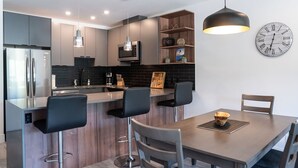 A modern kitchen with a central island, bar stools, a dining table, and a clock on the wall. - Alpine Haven by Revelstoke Vacations (Revelstoke)