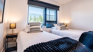 A hotel room with two beds, a nightstand with a lamp, a window with a view of trees and mountains, and a sign on the window. - Alpine Haven by Revelstoke Vacations (Revelstoke)