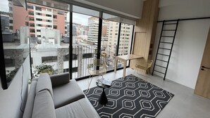 A modern living room with a grey sofa, a patterned rug, and a view of a cityscape through large windows. - Modern studio with parking - Murphy bed (Quito)