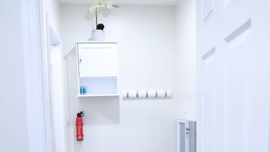 A narrow hallway with a white door on the right, a white shelf with a vase of flowers, and a red fire extinguisher on the left. - My Cozy-Snug Place - 1Brd - 8 mins to EWR (Elizabethport, NJ)