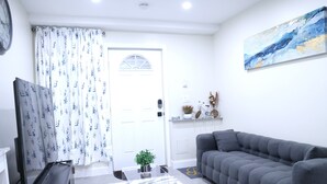 A living room with a grey sofa, a TV, a white door, and a painting on the wall. - My Cozy-Snug Place - 1Brd - 8 mins to EWR (Elizabethport, NJ)