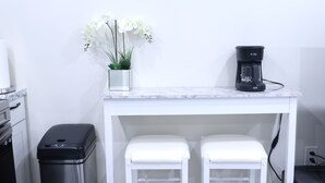 A kitchen with a white countertop, two white stools, a trash bin, a coffee maker, and a floral painting on the wall. - My Cozy-Snug Place - 1Brd - 8 mins to EWR (Elizabethport, NJ)