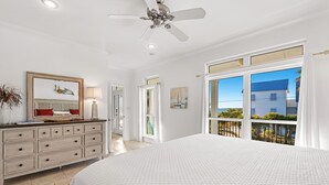 A bedroom with a large bed, a ceiling fan, a dresser, and a window with a view of buildings and greenery.