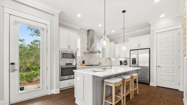 A modern kitchen with white cabinetry, a large island with stools, stainless steel appliances, and a view of greenery through a sliding door.
