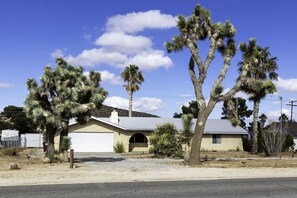 A single-story house with a garage, surrounded by desert vegetation, including a prominent Joshua tree. - Sky Harbor House - Hot Tub + Great Backyard Space (Yucca Valley)