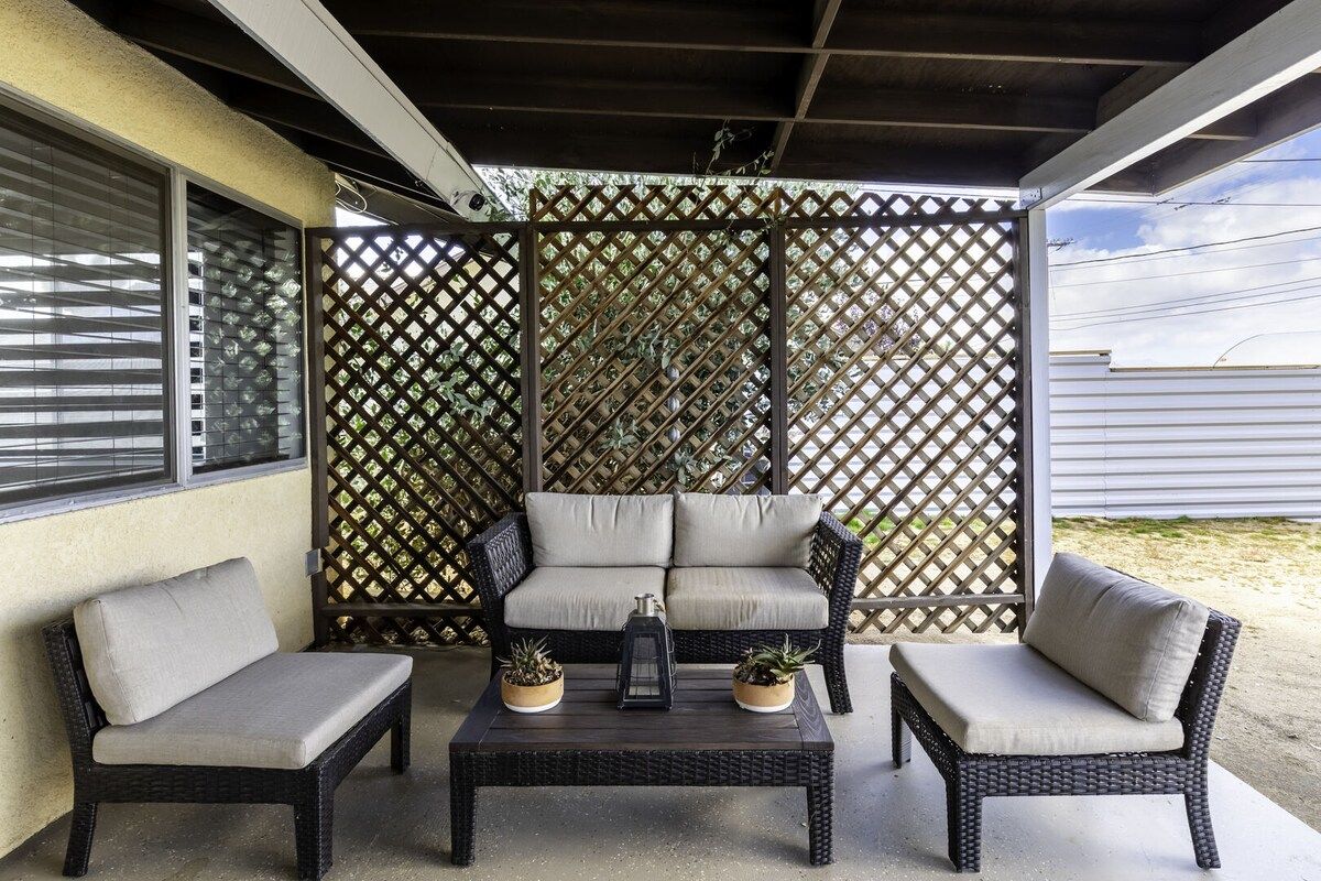 Un patio techado con un conjunto de sofá de mimbre, dos mesas auxiliares con cojines y plantas en macetas.