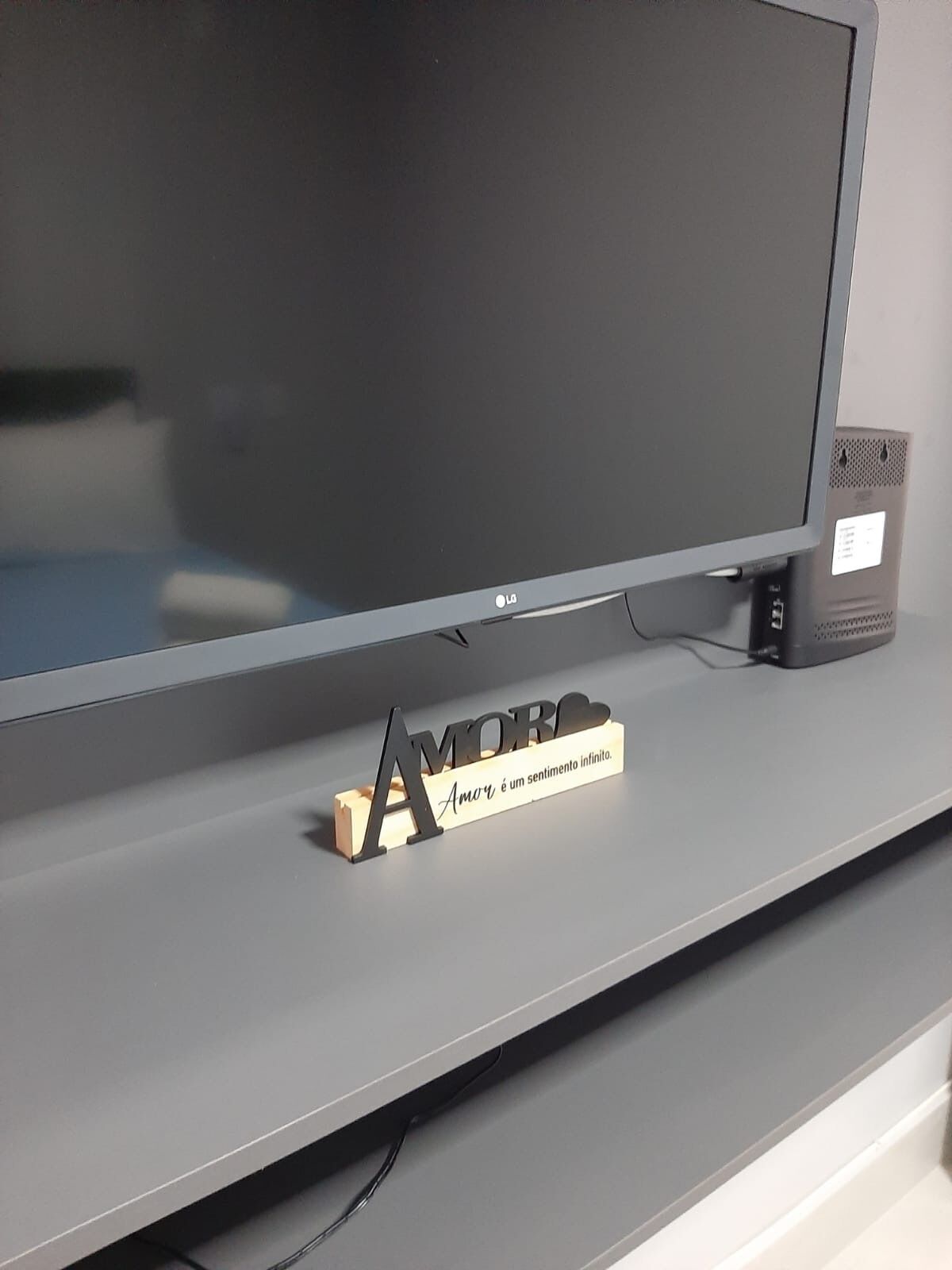 Un téléviseur à écran plat est posé sur un meuble de divertissement gris, accompagné d’un petit haut-parleur et d’une plaque en bois sur laquelle on peut lire : « L’amour est un sentiment infini. »