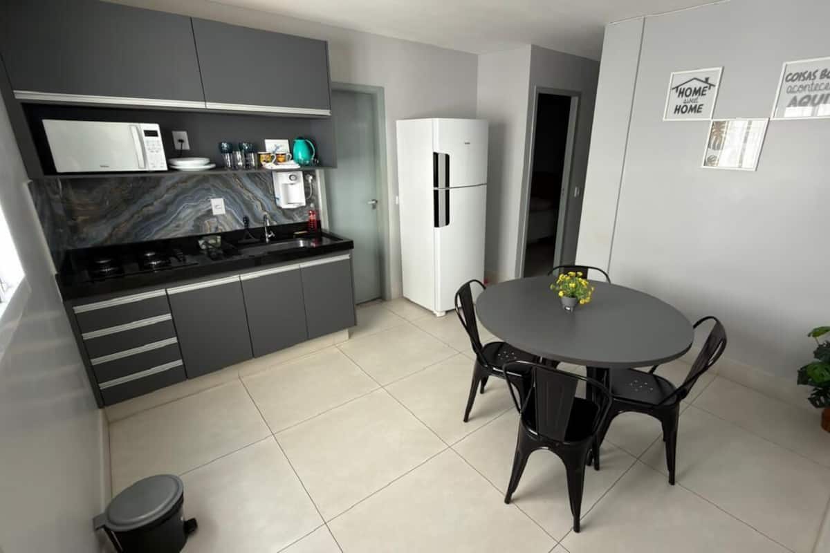 A modern kitchen with grey cabinets, a black countertop, and a white refrigerator. There is a dining table with chairs and a small potted plant.