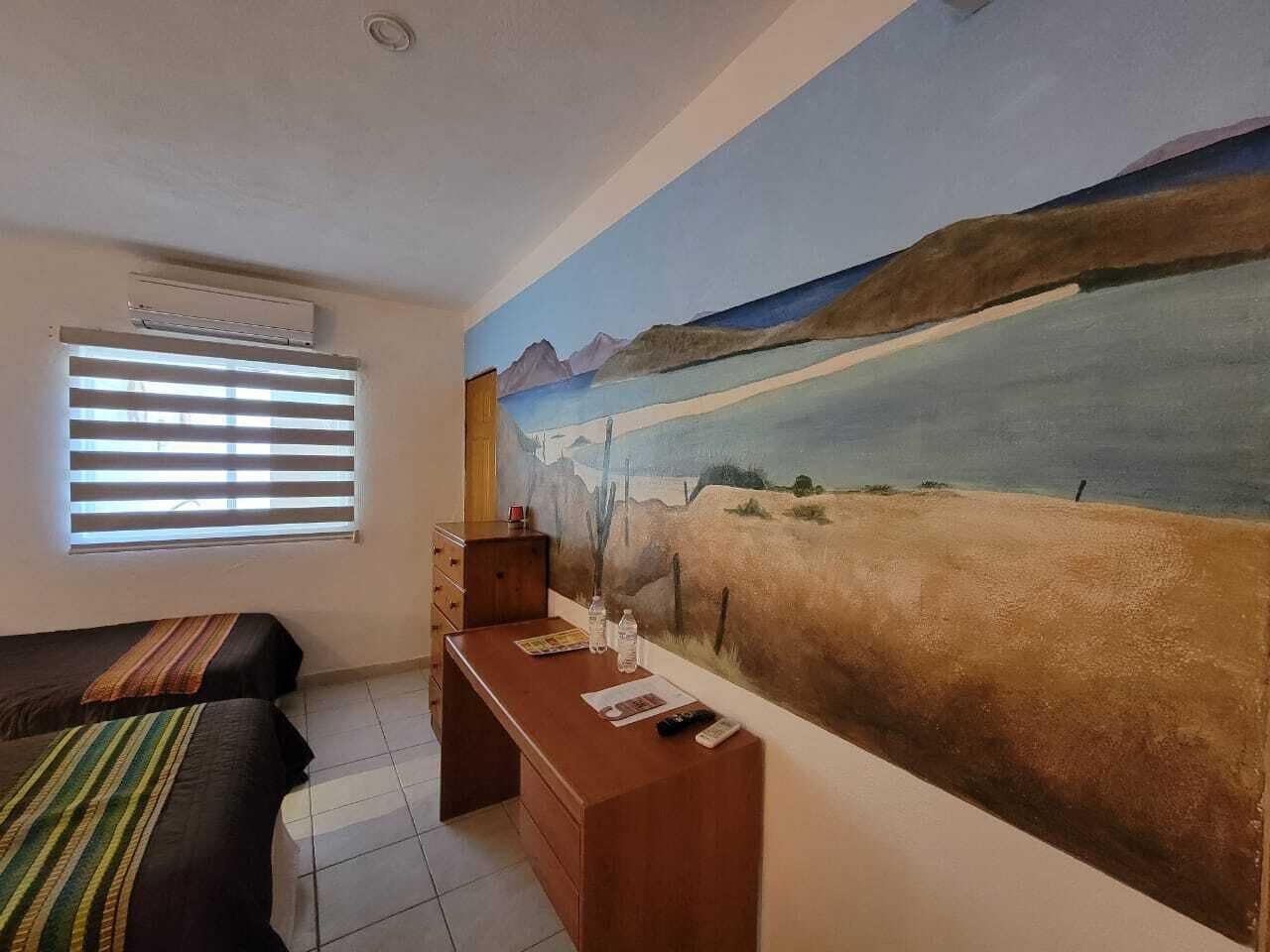 Ett rum med två sängar, ett skrivbord, ett fönster med persienner och en stor landskapsmålning på väggen.