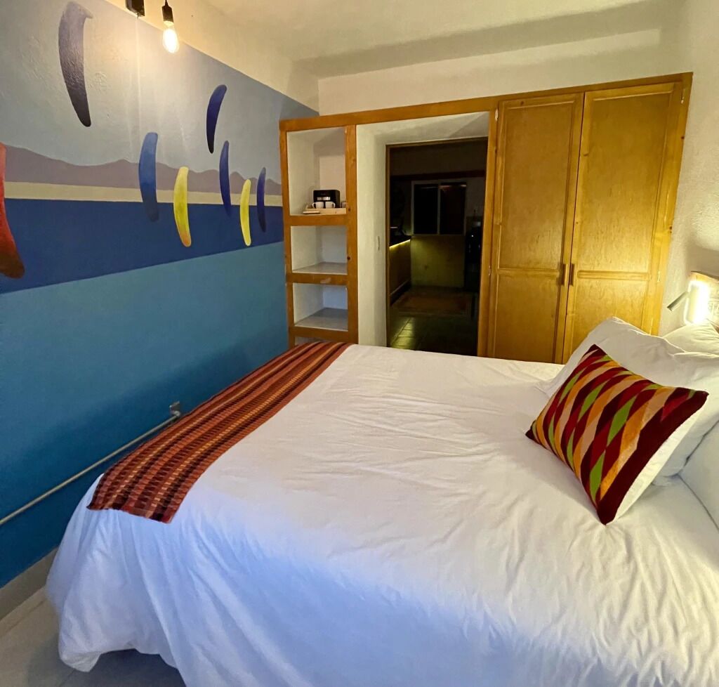 A bedroom with a bed, a colorful wall mural, a wooden wardrobe, and a small shelf with a coffee maker.