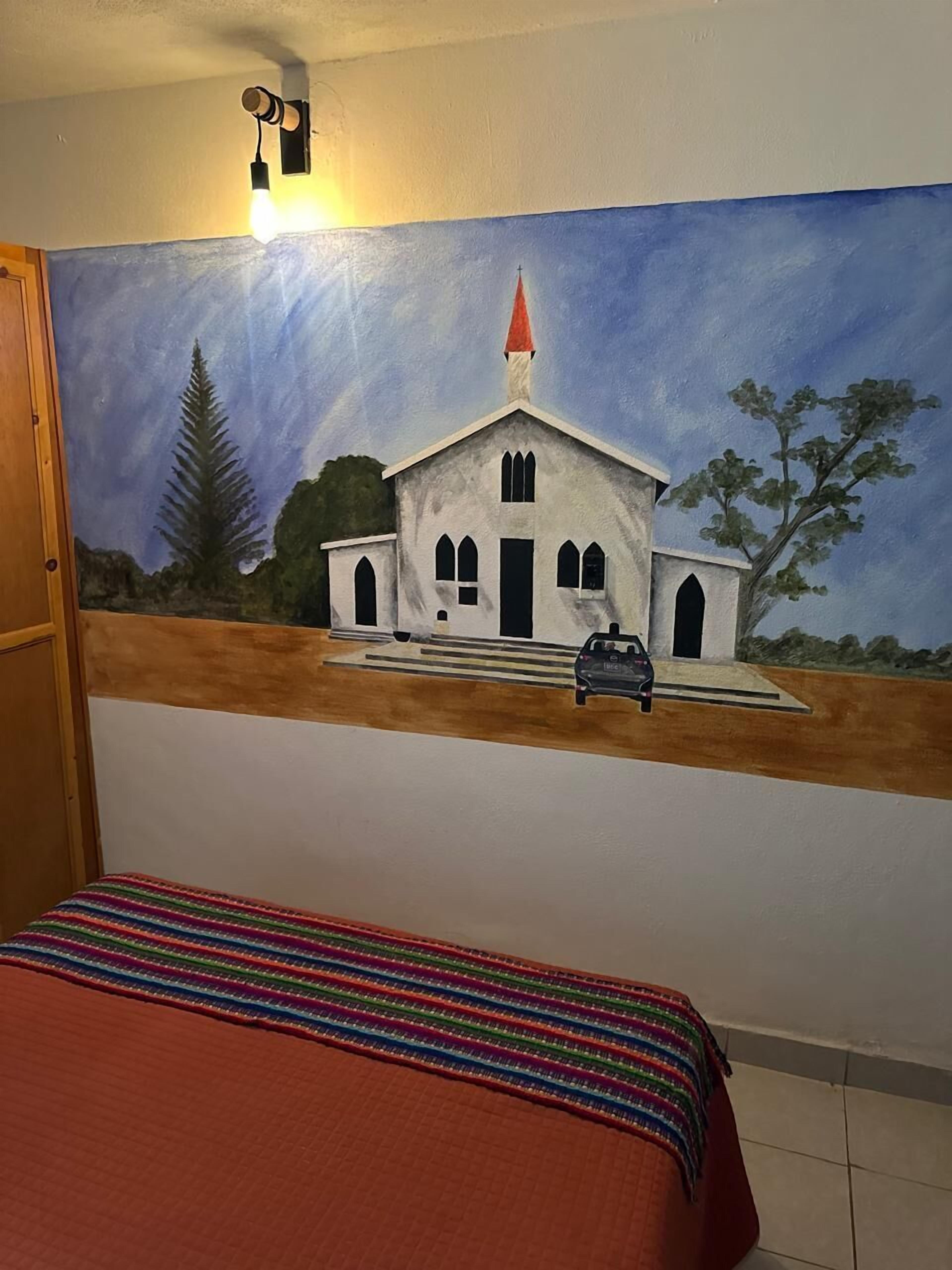 Quarto com uma cama, um quadro de uma igreja e uma luz noturna.