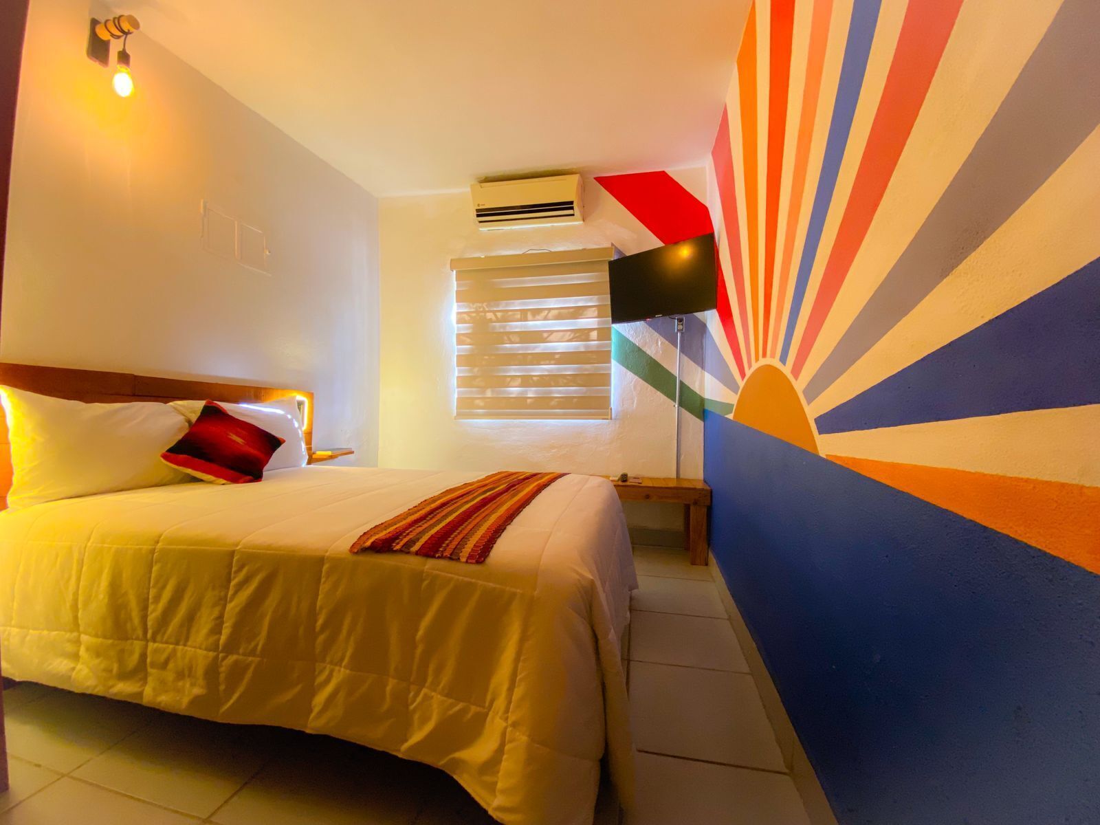 Una habitación de hotel con una cama, una mesita de noche, un equipo de aire acondicionado, un televisor montado en la pared y una ventana con persianas.