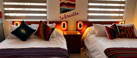 A hotel room with two beds, each with colorful pillows and a striped blanket. A wall mural reads "TAKE A MOMENT to Breathe."