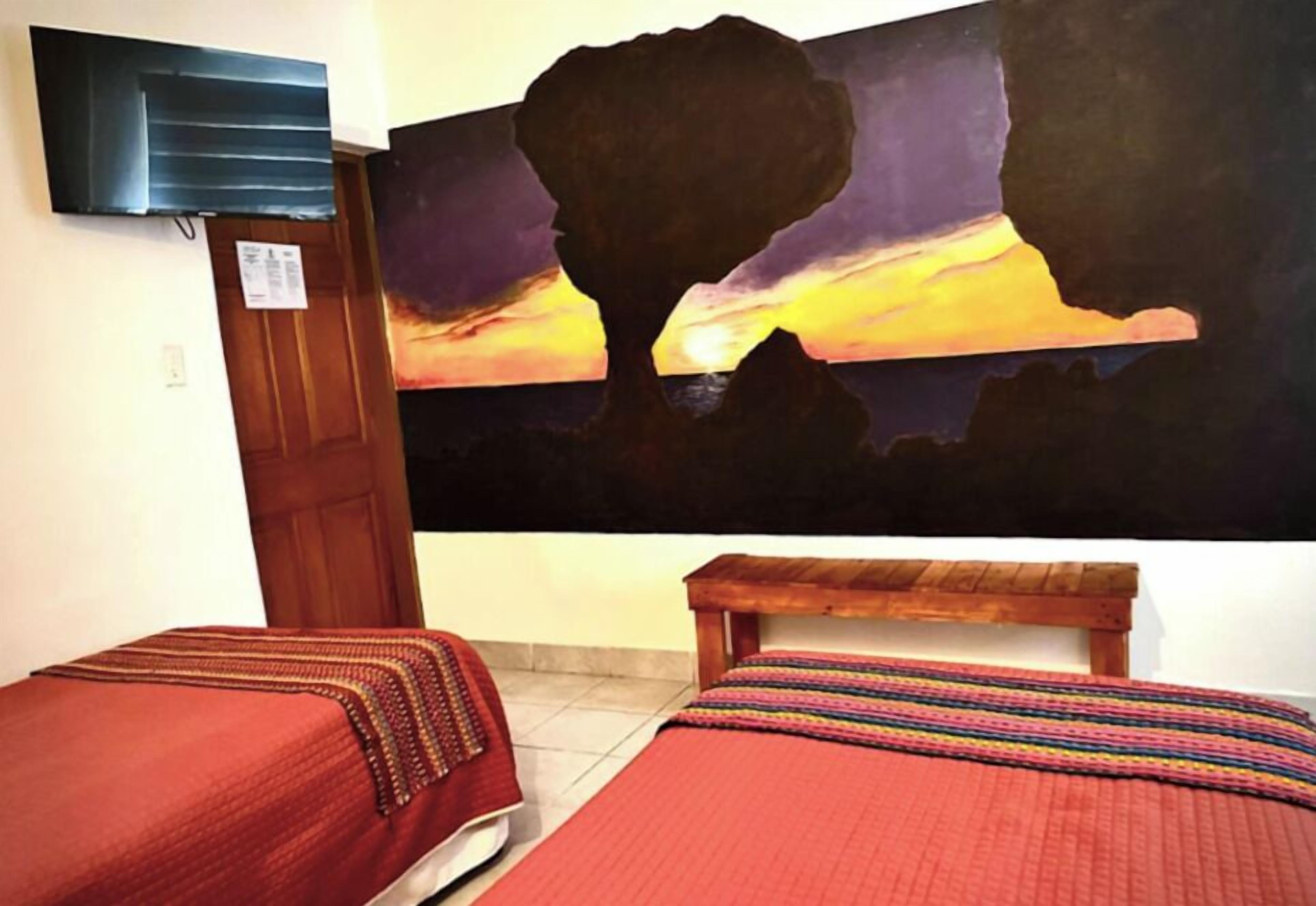 Une chambre avec deux lits, un tableau représentant la silhouette d’un arbre se détachant sur un coucher de soleil, et un téléviseur fixé au mur.