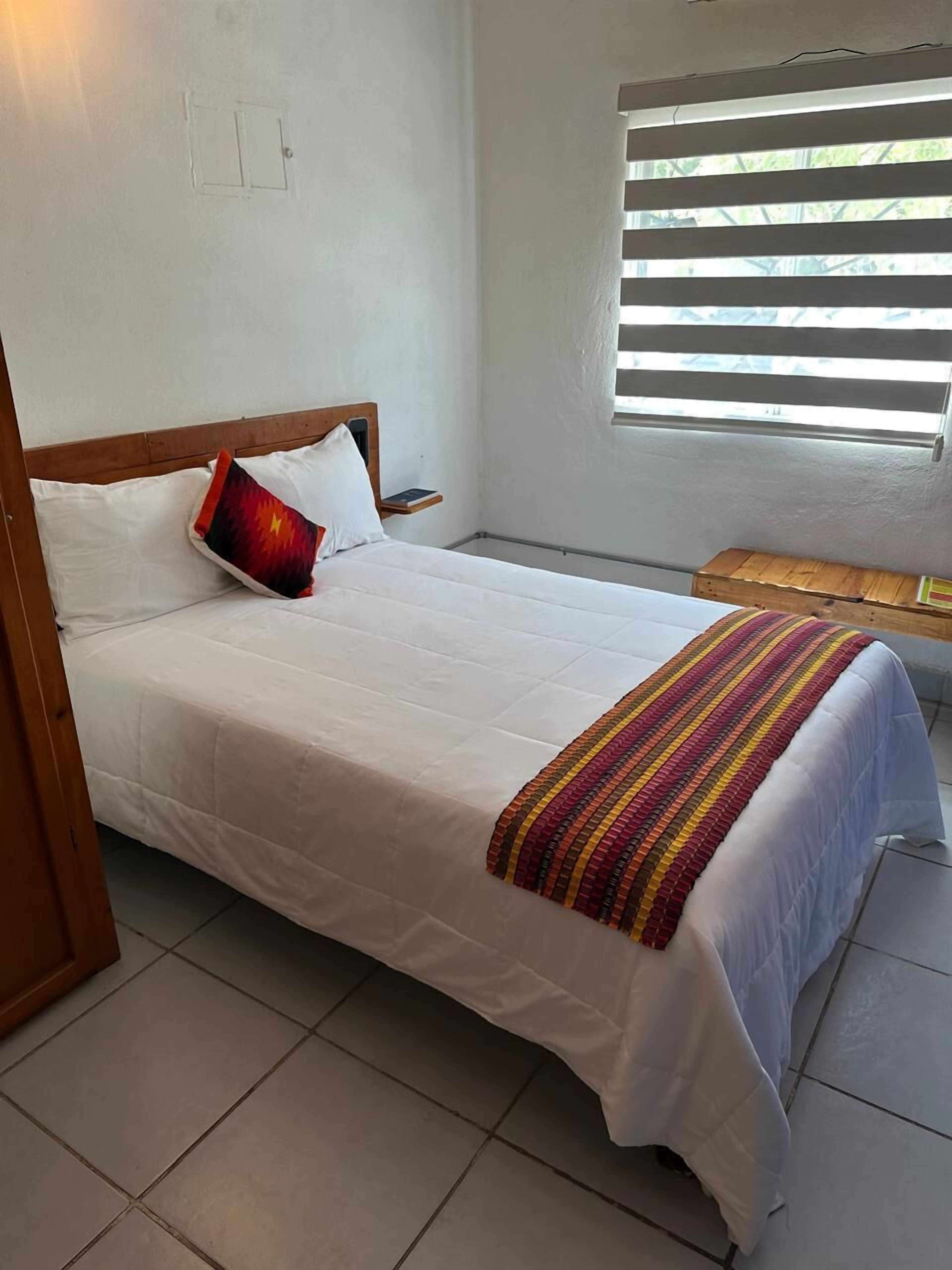 A bed with white bedding and a colorful striped blanket, a wooden headboard, and a window with blinds.