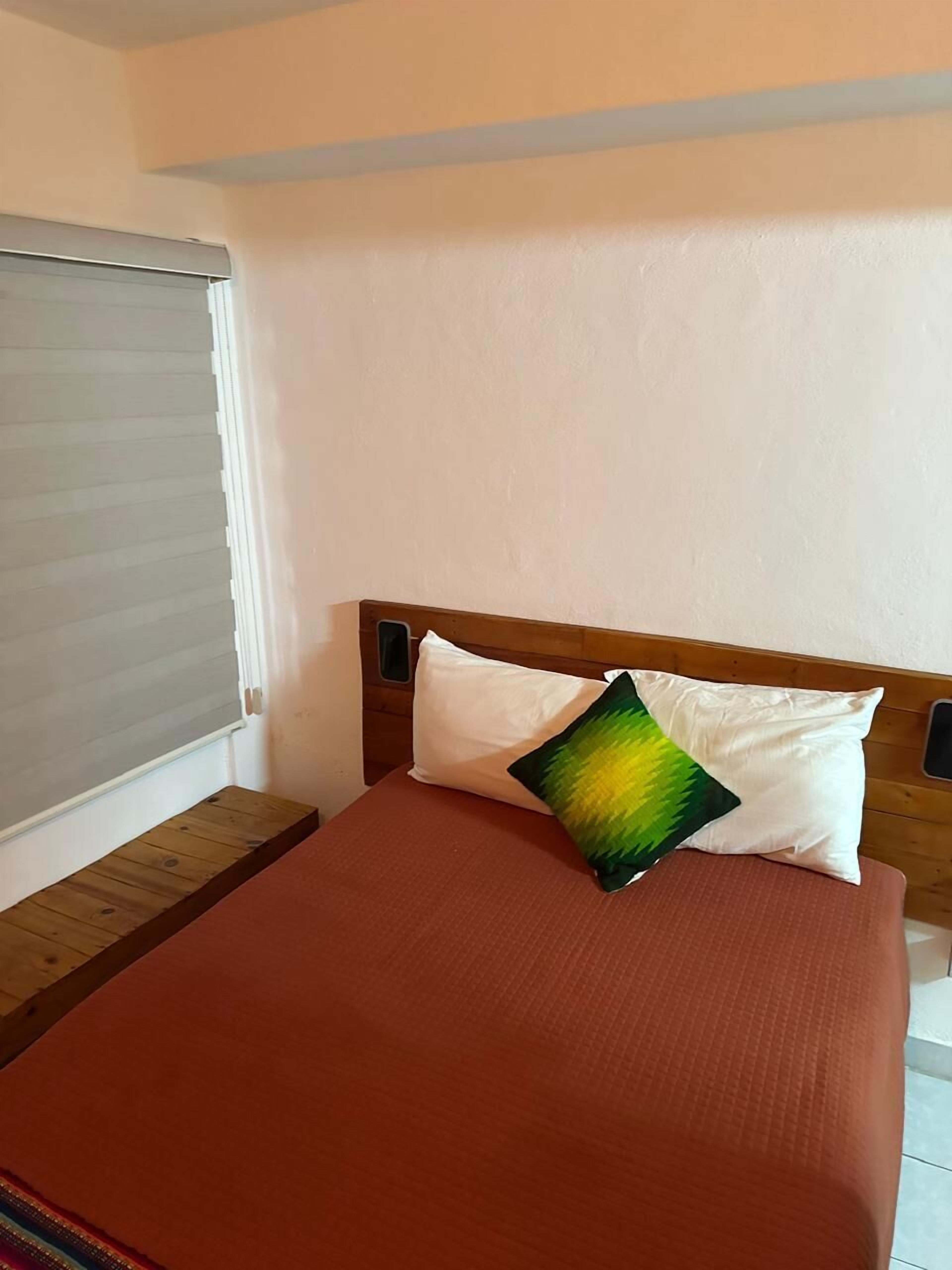 Una cama con cabecera de madera, una colcha roja y dos almohadas, una blanca y otra multicolor.