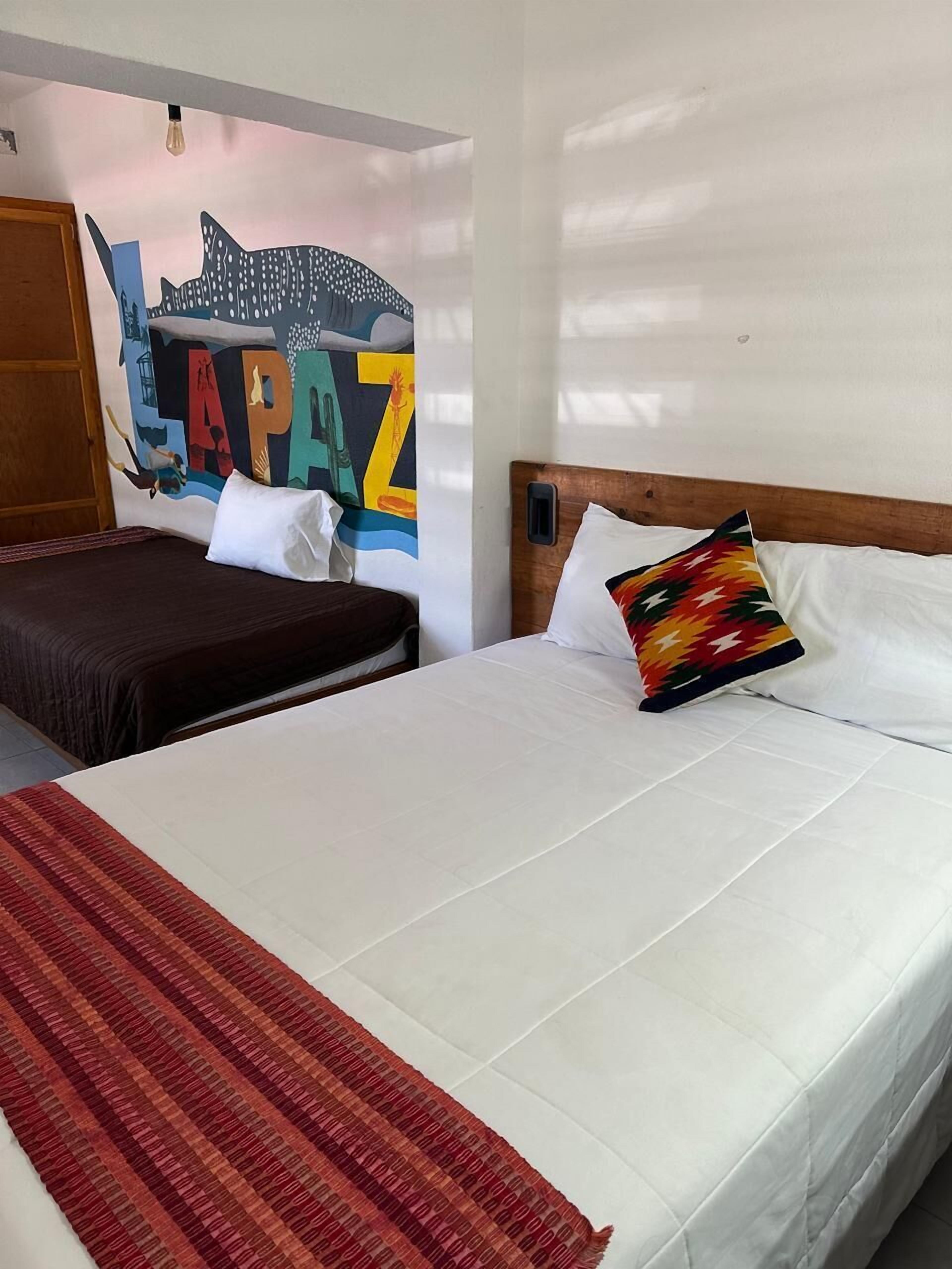 Una habitación de hotel con una cama grande, una cama más pequeña, un mural colorido en la pared y una alfombra roja a rayas.