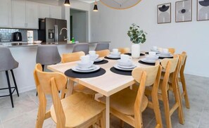 A modern dining area with a wooden table set for 10 people, surrounded by grey chairs. A kitchen with modern appliances is visible in the background. - 3 BR, 2.5 BA Villa | Sleeps 10! (Davenport)