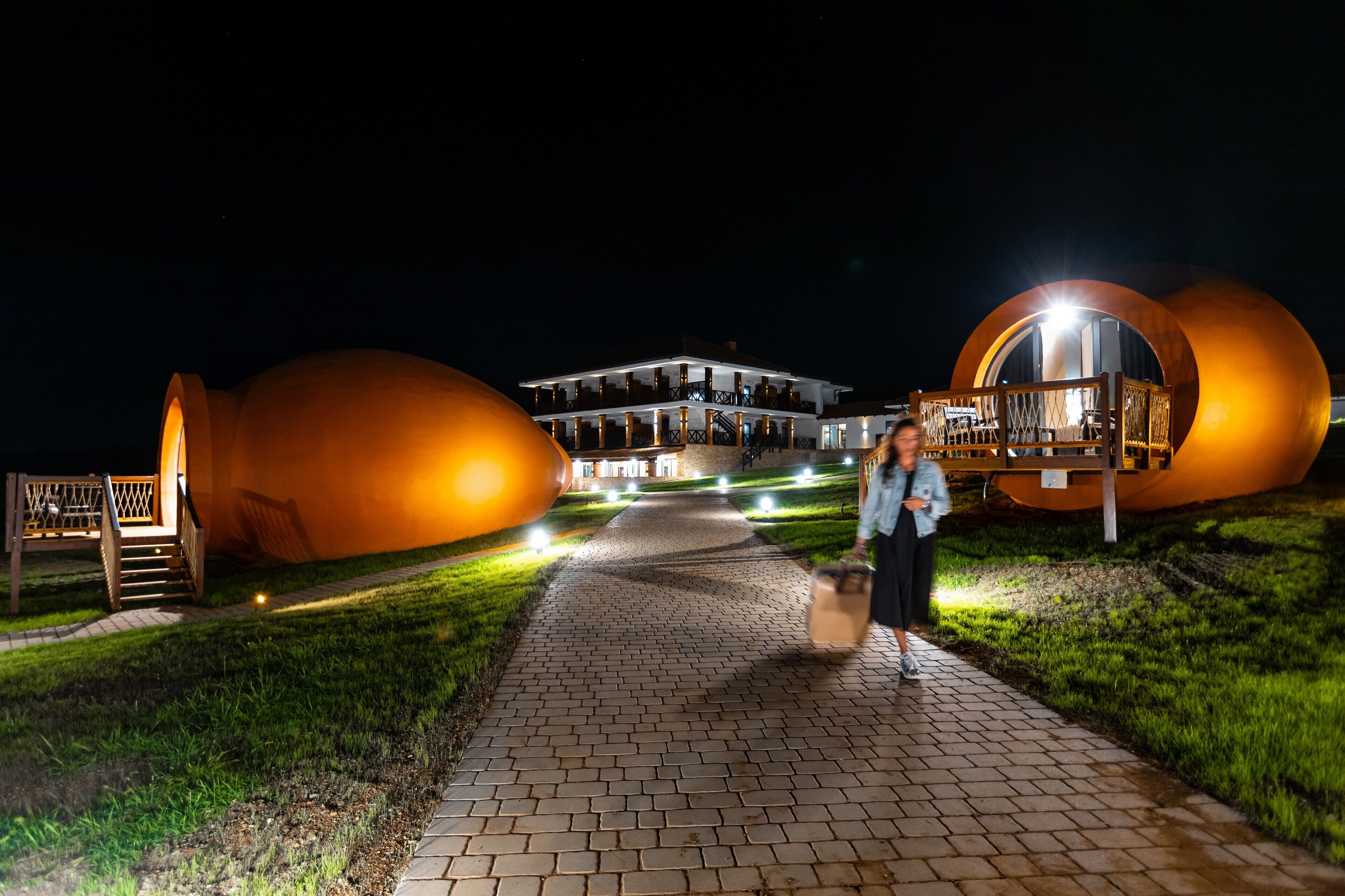 A pathway leading to a building with a unique circular design, a well-lit entrance, and a visible outdoor seating area.
