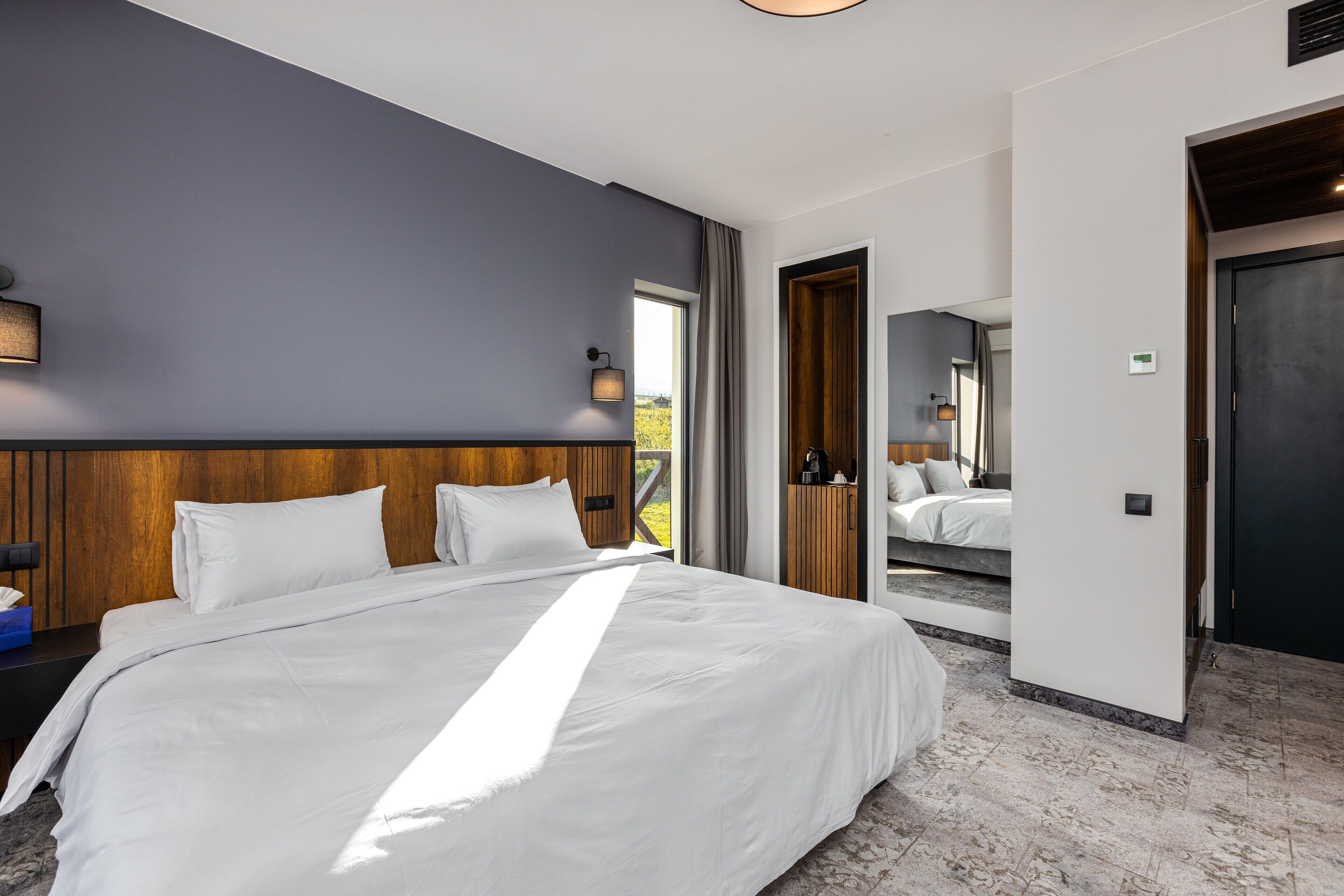 A modern hotel room with a large bed, a wooden headboard, a nightstand, a bedside table, a lamp, and a view of the outdoors.