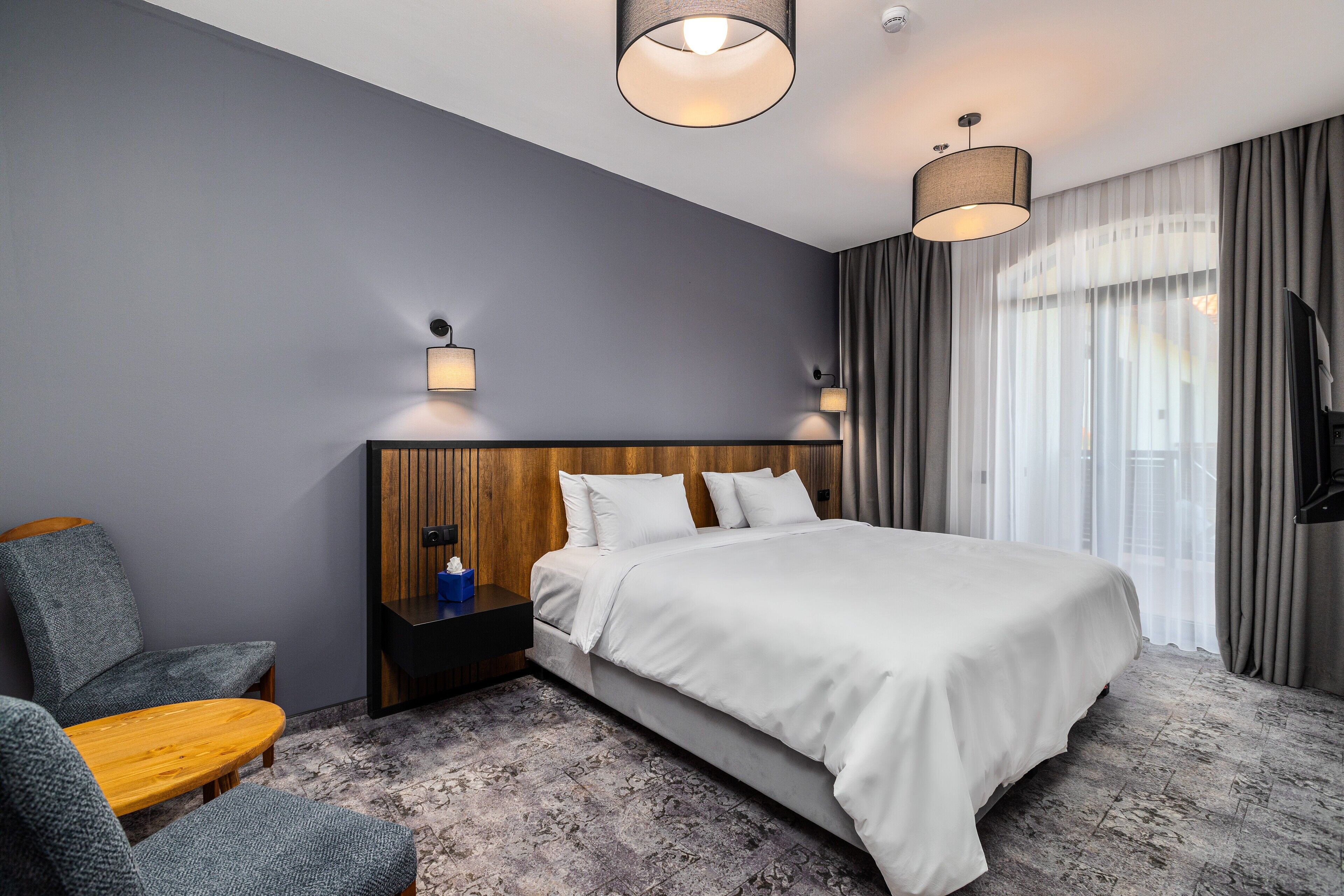 A modern hotel room with a large bed, a wooden headboard, a nightstand, a seating area with a chair and table, and a TV.