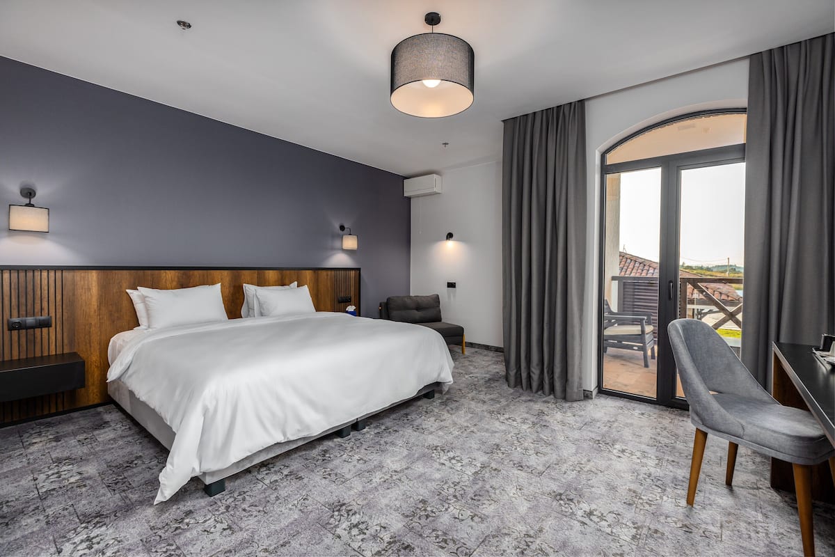 A modern hotel room with a large bed, a desk, and a view of the outdoors through a sliding door.