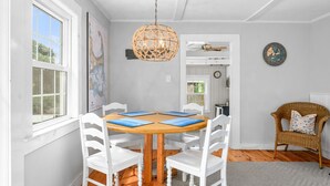 Un coin repas avec une table ronde en bois, des chaises blanches, un luminaire suspendu en rotin, une carte accrochée au mur et un fauteuil en osier avec un coussin.