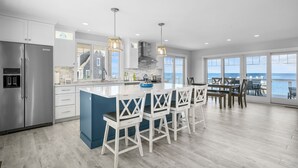 A modern kitchen with a central island, bar stools, and a view of the ocean through large windows. - Oceanfront Cape Escape with Panoramic Bay Views (East Sandwich)