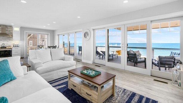 A spacious living room with a large window overlooking the ocean, a kitchen area, and a balcony with outdoor seating. - Oceanfront Cape Escape with Panoramic Bay Views (East Sandwich)