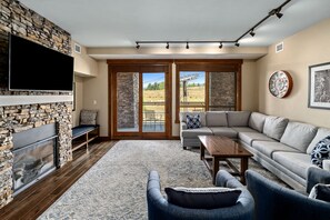 A living room with a stone fireplace, a flat-screen TV, a grey sofa, a coffee table, and a view of a grassy area through glass doors. - Lupine Lodge, AC! Private Deck with Views! Pool/Hot Tubs, Amenities Galore! (Steamboat Springs)