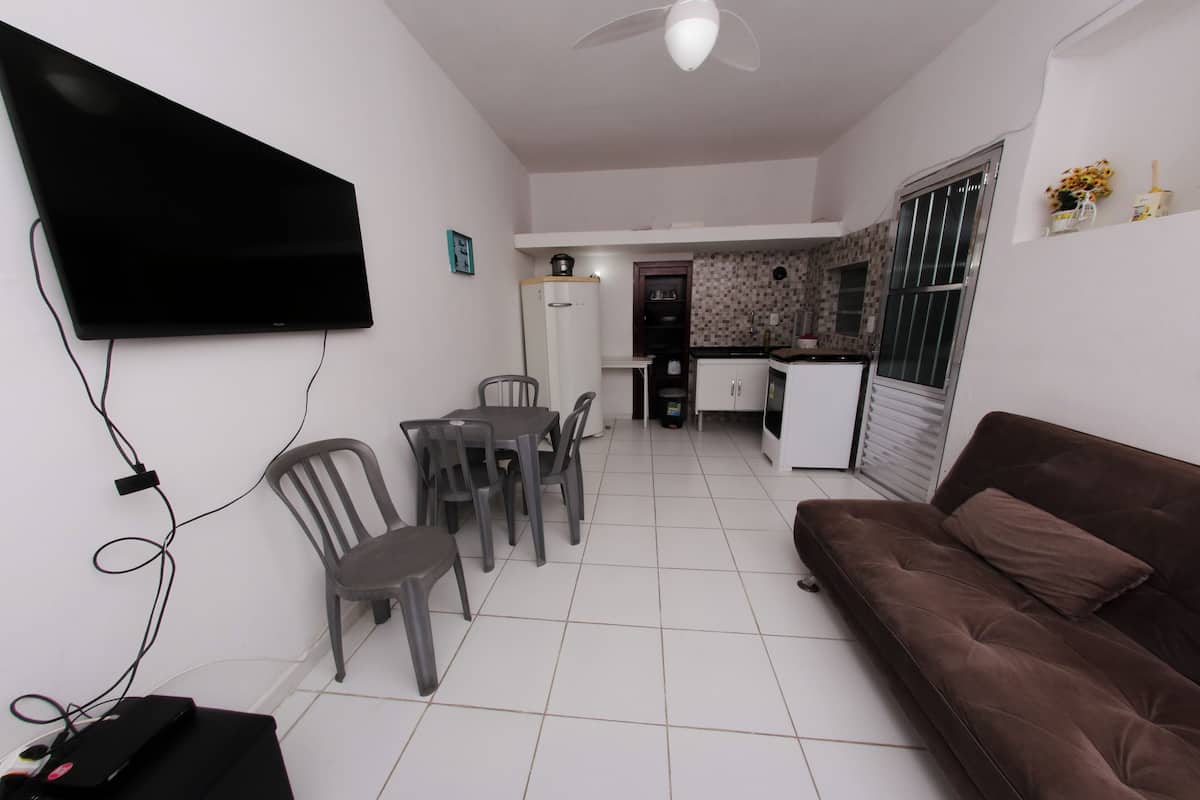 A living room with a flat-screen TV mounted on the wall, a brown sofa, and a dining area with plastic chairs and a table.
