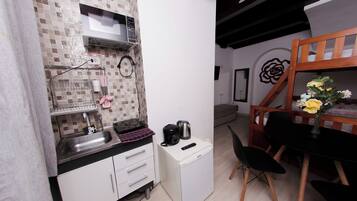 A compact kitchen with a microwave, kettle, and coffee maker on a small white cabinet. There is a sink with a faucet, and a wooden chair and table with a vase of flowers.