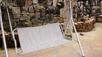 Una silla colgante de metal con un cojín gris frente a un muro de piedra.